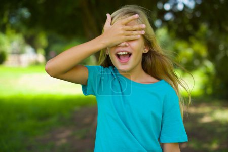 Porträt eines schönen kaukasischen kleinen Mädchens mit blauem T-Shirt, das draußen im Park steht und sein Gesicht verdeckt und lächelt