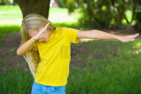Photo for Photo of funky caucasian little girl wearing yellow t-shirt standing outdoors show disco move dab - Royalty Free Image