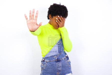 Photo for Young African American woman with short hair wearing denim overall against white wall covers eyes with palm and doing stop gesture, tries to hide. Don't look at me, I don't want to see, feels ashamed or scared. - Royalty Free Image