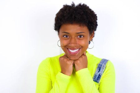 Foto de Mujer afroamericana joven satisfecha con el pelo corto que usa denim en general contra la pared blanca toca la barbilla con ambas manos, sonríe agradablemente, se regocija buen día con el amante - Imagen libre de derechos