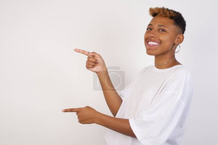 Téléchargez les photos : Positif attrayant jeune femme afro-américaine portant des points de t-shirt blanc de côté avec une expression joyeuse et surprise avec la bouche ouverte, montre quelque chose d'étonnant debout à l'intérieur. Concept de publicité. - en image libre de droit