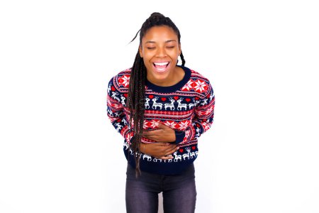 Téléchargez les photos : Femme afro-américaine portant un pull de Noël contre un mur blanc garde les mains croisées, rit à haute voix à la bonne blague, porte des vêtements décontractés. - en image libre de droit