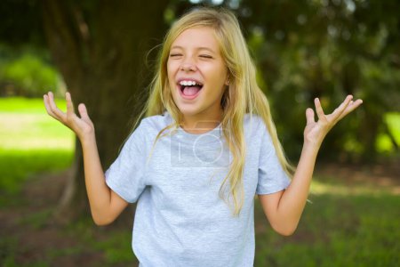 Photo for Crazy outraged caucasian little kid girl wearing white t-shirt standing outdoor in the park screams loudly and gestures angrily yells furiously. Negative human emotions feelings concept - Royalty Free Image