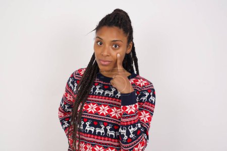 Foto de Mujer afroamericana vistiendo suéter de Navidad contra pared blanca mirando, observando, vigilando un objeto en frente, o mirando hacia fuera para algo. - Imagen libre de derechos