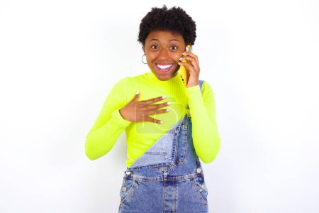Foto de Sonriente joven mujer afroamericana con el pelo corto con denim en general contra las conversaciones de pared blanca a través del teléfono celular, disfruta de una agradable gran conversación. Personas, tecnología, concepto de comunicación - Imagen libre de derechos