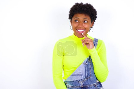 Photo for Young African American woman with short hair wearing denim overall against white with thoughtful expression, looks to the camera, keeps hand near face, biting a finger thinks about something pleasant. - Royalty Free Image
