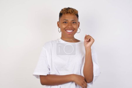 Téléchargez les photos : Image d'une jolie jeune femme afro-américaine joyeuse portant un t-shirt blanc debout contre un mur gris avec la main près du visage. Regarder avec joie l'expression à la caméra après avoir écouté les bonnes nouvelles. Fille confiante. - en image libre de droit