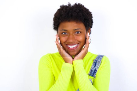 Foto de Feliz joven mujer afroamericana con el pelo corto con denim en general contra la pared blanca toca ambas mejillas suavemente, tiene una sonrisa tierna, muestra dientes blancos, mira positivamente directamente a la cámara, - Imagen libre de derechos