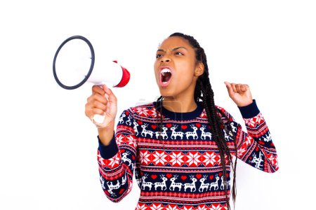 Foto de Mujer afroamericana vistiendo suéter de Navidad contra la pared blanca se comunica gritando en voz alta sosteniendo un megáfono, expresando el éxito y el concepto positivo, idea de marketing o ventas. - Imagen libre de derechos