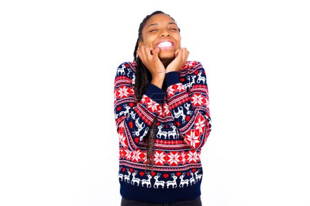 Téléchargez les photos : Portrait de femme afro-américaine portant un pull de Noël contre un mur blanc submergé, exprimant l'excitation et le bonheur avec les yeux fermés et les mains près du visage. - en image libre de droit