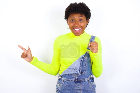 Foto de Joven mujer afroamericana con el pelo corto con denim en general contra los puntos de la pared blanca en el espacio vacío sosteniendo el puño hacia arriba, gesto ganador. - Imagen libre de derechos