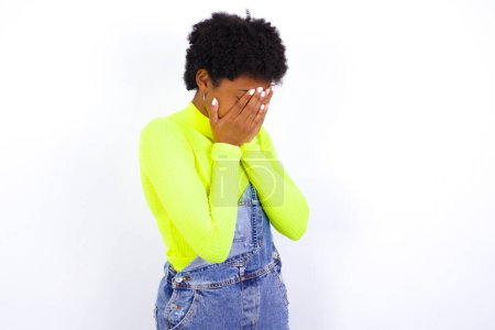 Téléchargez les photos : Triste jeune femme afro-américaine aux cheveux courts portant globalement du denim contre le mur blanc couvrant le visage avec les mains et pleurant. - en image libre de droit