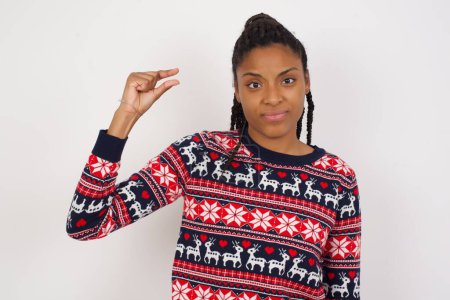 Foto de Mujer afroamericana molesta usando suéter de Navidad contra formas blancas de la pared poco gesto con la mano demuestra algo muy pequeño tamaño. No mucho. - Imagen libre de derechos