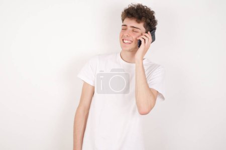 Téléchargez les photos : Drôle beau jeune homme sur fond blanc rit heureusement, a conversation téléphonique, étant amusé par un ami, ferme les yeux. - en image libre de droit