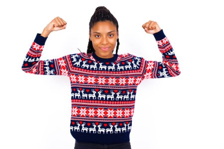 Photo for Waist up shot of African American woman wearing Christmas sweater against white wall raises arms to show muscles feels confident in victory, looks strong and independent, smiles positively at camera. Sport concept. - Royalty Free Image