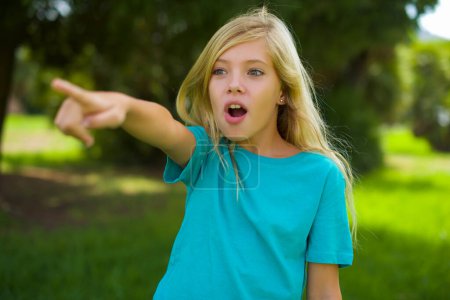 Porträt eines schönen kaukasischen kleinen Mädchens mit blauem T-Shirt, das draußen im Park steht und mit dem Finger zeigt