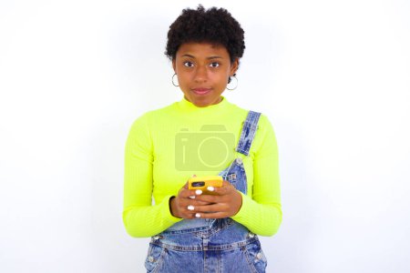 Foto de Joven mujer afroamericana con el pelo corto con denim en general contra la pared blanca Simular espacio de copia. Usando el teléfono móvil, escribiendo el mensaje sms - Imagen libre de derechos