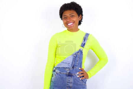 Foto de Estudio disparo de alegre joven afroamericana mujer con el pelo corto con denim en general contra blanco mantiene la mano en la cadera, sonríe ampliamente. - Imagen libre de derechos