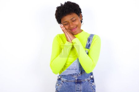 Foto de Joven mujer afroamericana con el pelo corto con denim en general contra la pared blanca se apoya en las palmas prensadas cierra los ojos y tiene agradables sueños sonrisa sobre algo - Imagen libre de derechos