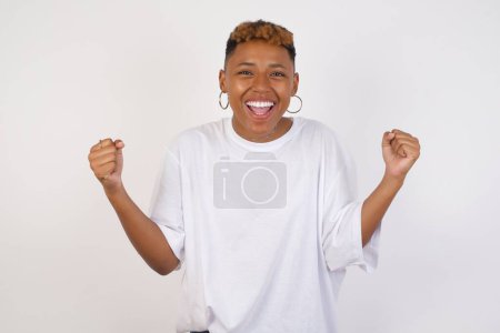 Foto de Retrato de cintura hacia arriba de una joven afroamericana decidida y con éxito fuerte que usa una camiseta blanca ganadora en una camiseta casual levantando brazos, apretando puños, exclamando con alegría y emoción. Concepto de victoria, éxito y logro. - Imagen libre de derechos