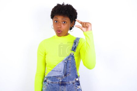 Photo for Shocked young African American woman with short hair wearing denim overall against white shows something little with hands, demonstrates size, opens mouth from surprise. Measurement concept. - Royalty Free Image
