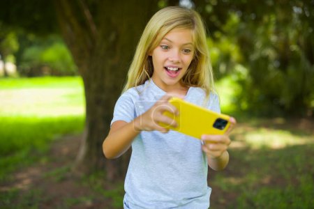 Portrait of caucasian little kid girl wearing white t-shirt standing outdoor in the park playing game on smartphone