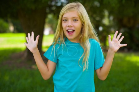 Téléchargez les photos : Portrait de belle petite fille caucasienne portant t-shirt bleu debout en plein air dans le parc - en image libre de droit