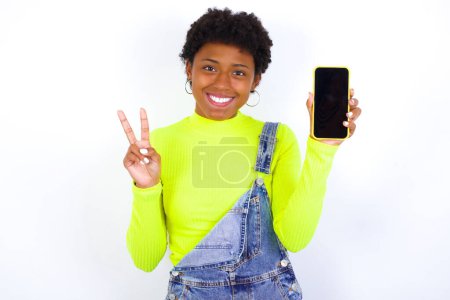 Foto de Joven mujer afroamericana con el pelo corto usando denim en general contra la pared blanca sosteniendo teléfono inteligente y mostrando señal de paz - Imagen libre de derechos