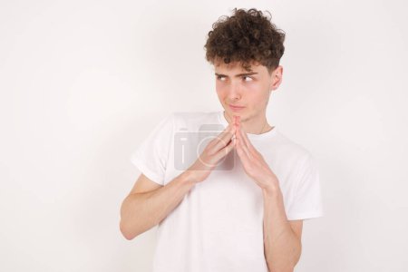 Photo for Handsome young man over white background steeples fingers and looks mysterious aside has great evil plan in mind - Royalty Free Image