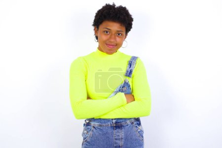 Photo for Self confident serious calm young African American woman with short hair wearing denim overall against white wall stands with arms folded. Shows professional vibe stands in assertive pose. - Royalty Free Image