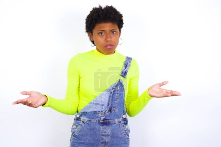 Photo for Young African American woman with short hair wearing denim overall against white wall looks uncertain shrugs shoulders. - Royalty Free Image