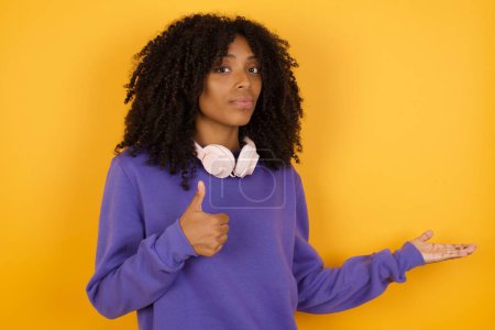 Téléchargez les photos : Portrait de jeune femme afro-américaine expressive avec écouteurs sur fond jaune pointant vers l'espace vide - en image libre de droit