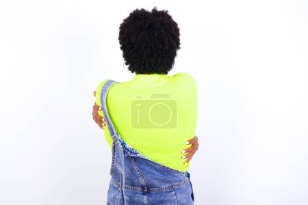 Foto de Joven mujer afroamericana con el pelo corto con denim en general contra la pared blanca abrazándose feliz y positivo desde atrás. Amor propio y cuidado personal. - Imagen libre de derechos