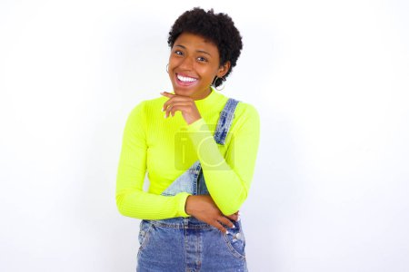 Photo for Young African American woman with short hair wearing denim overall against white wall laughs happily keeps hand on chin expresses positive emotions smiles broadly has carefree expression - Royalty Free Image