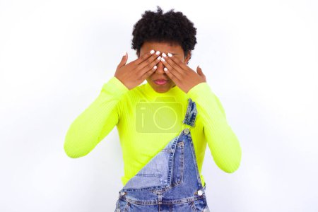 Foto de Joven mujer afroamericana con el pelo corto con denim en general contra la pared blanca que cubre los ojos con ambas manos, no quiere ver nada o sentirse avergonzado. Reacciones de sentimientos humanos. - Imagen libre de derechos