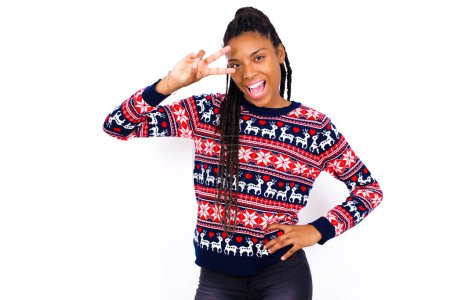 Téléchargez les photos : Femme afro-américaine portant un pull de Noël contre un mur blanc faisant signe en V près des yeux. Loisirs, coquettes, célébration et concept de flirt. - en image libre de droit