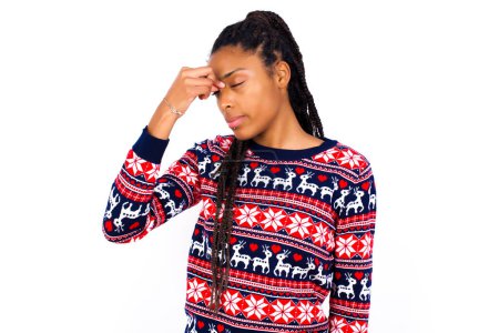 Foto de Mujer afroamericana muy molesta vistiendo suéter de Navidad contra pared blanca tocando la nariz entre los ojos cerrados, quiere llorar, tener una relación estresante o tener problemas con el trabajo - Imagen libre de derechos