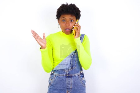 Téléchargez les photos : Jeune afro-américaine aux cheveux courts portant globalement du denim contre un mur blanc parlant au téléphone stressé avec la main sur le visage, choqué par la honte et le visage surprise, en colère et frustré. Peur et contrariété par erreur. - en image libre de droit