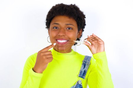 Téléchargez les photos : Jeune afro-américaine aux cheveux courts portant globalement du denim contre du blanc tenant un aligneur invisible et pointant vers ses dents droites parfaites. Concept de soins dentaires et de confiance. - en image libre de droit