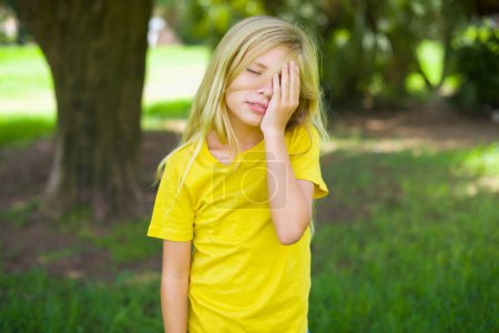 Téléchargez les photos : Petite fille caucasienne portant un t-shirt jaune debout à l'extérieur avec une expression triste couvrant le visage avec les mains tout en pleurant. Concept de dépression. - en image libre de droit
