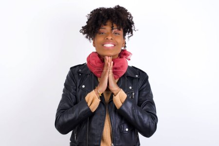 Photo for Young beautiful African American woman wearing biker jacket against white wall keeps palms together, has pleased expression. Glad attractive male makes request, pleads for mercy. Hopeful young adult. - Royalty Free Image