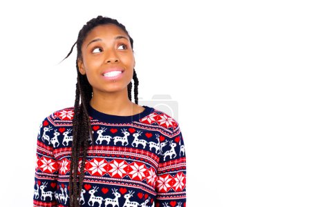 Téléchargez les photos : Oups ! Portrait de femme afro-américaine portant un pull de Noël contre un mur blanc serre les dents et regarde de côté confusément, réalise sa mauvaise erreur, - en image libre de droit