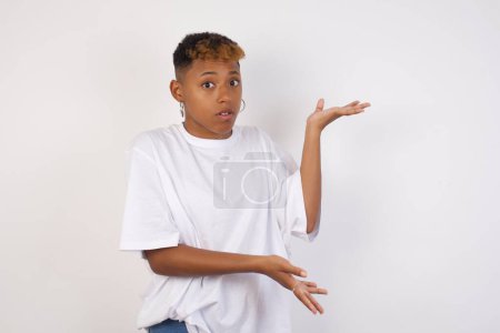 Téléchargez les photos : Belle femme afro-américaine portant un t-shirt blanc pointant de côté avec les deux mains montrant quelque chose d'étrange et disant : Je ne sais pas ce que c'est. Debout sur fond gris. Concept de publicité. - en image libre de droit