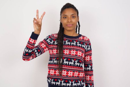 Foto de Mujer afroamericana vistiendo jersey de Navidad contra pared blanca mostrando y apuntando hacia arriba con los dedos número dos mientras sonríe confiada y feliz. - Imagen libre de derechos