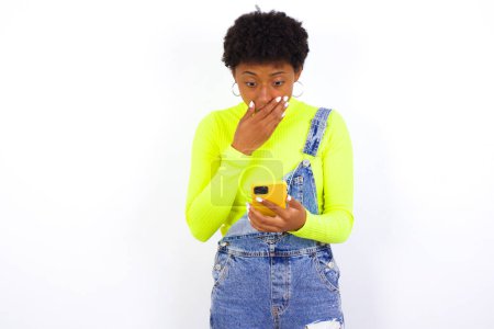Photo for Young African American woman with short hair wearing denim overall against white being deeply surprised, stares at smartphone display, reads shocking news on website, Omg, its horrible! - Royalty Free Image