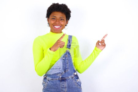 Foto de Joven mujer afroamericana positiva con el pelo corto usando denim en general contra la pared blanca con sonrisa radiante señalando con dos dedos y mirando en el espacio de copia vacío. Concepto de publicidad. - Imagen libre de derechos