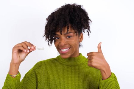 Téléchargez les photos : Jeune belle femme afro-américaine portant un pull tricoté vert contre un mur blanc, tenant des bretelles d'alignement invisibles et levant le pouce vers le haut. Recommandation et concept de soins dentaires. - en image libre de droit