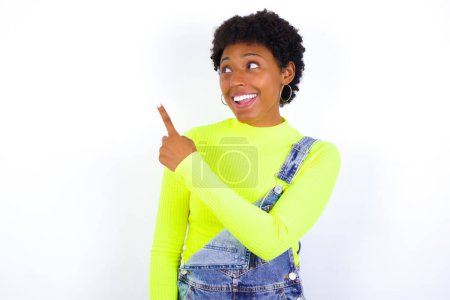 Téléchargez les photos : Jeune afro-américaine avec les cheveux courts portant le denim dans l'ensemble contre le mur blanc joyeuse démonstration de l'espace de copie regarder nouveauté - en image libre de droit