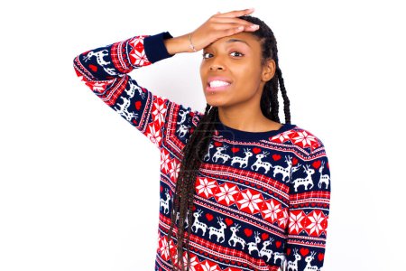 Téléchargez les photos : Qu'est-ce que j'ai fait ? Femme afro-américaine portant un pull de Noël contre un mur blanc tenant la main sur le front avec une expression de peur et de regret. - en image libre de droit