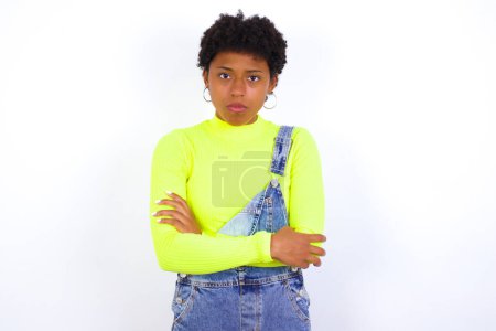Foto de Foto de la joven mujer afroamericana enojada con el pelo corto que usa denim en general contra la cámara de aspecto de pared blanca. - Imagen libre de derechos